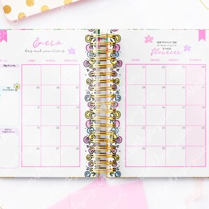 May include: Open planner with a gold spiral binding, featuring a pink and white calendar layout. The calendar pages are decorated with floral and whimsical designs, with text in Spanish. The planner is open to a January spread.
