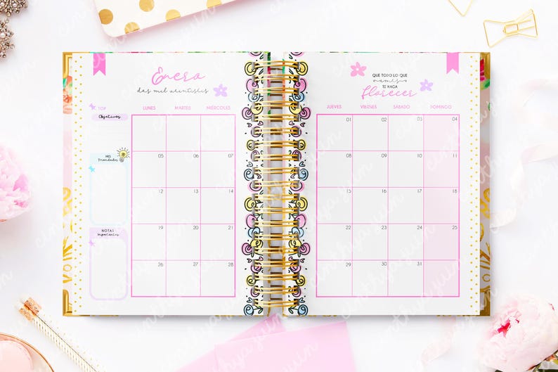 May include: Open planner with a gold spiral binding, featuring a pink and white calendar layout. The calendar pages are decorated with floral and whimsical designs, with text in Spanish. The planner is open to a January spread.