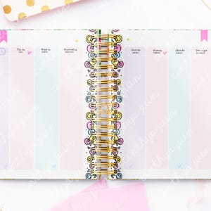 May include: Open planner with a gold spiral binding, featuring pastel-colored columns for each day of the week. The pages are decorated with floral and geometric patterns, and the text is in Spanish.