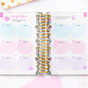 May include: Open notebook with a gold spiral binding, featuring pastel-colored monthly birthday tracking pages. The pages are decorated with whimsical illustrations and the Spanish text "cumplea&ntilde;os especiales".