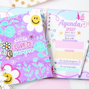 Puede incluir: Una agenda 2027 de espiral con portada morada, flores blancas y amarillas con caras sonrientes, dibujos lineales de flores en turquesa y el texto Florece siempre. La página abierta muestra Agenda 2027, Mereces todo lo bonito de la vida.