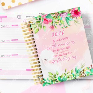 May include: Open spiral-bound planner with a pink cover featuring floral accents and the year 2026. The cover has text in Spanish. The open pages show a weekly layout with pink accents and gold spiral binding. A gold pen and other stationery items are in the background.