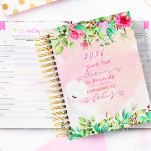 May include: A spiral-bound planner with a pink cover featuring the year 2026 and floral illustrations. The cover has a quote in Spanish. The open planner reveals lined pages with gold spiral binding and a gold-colored border.