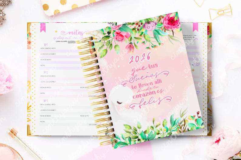 May include: A spiral-bound planner with a pink cover featuring the year 2026 and floral illustrations. The cover has a quote in Spanish. The open planner reveals lined pages with gold spiral binding and a gold-colored border.