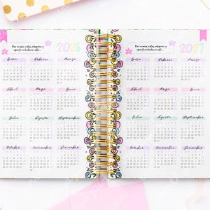 May include: Open planner with 2026 and 2027 calendars. The pages are white with pastel-colored month headings and gold spiral binding. The planner has a floral design on the edges and includes text in Spanish.