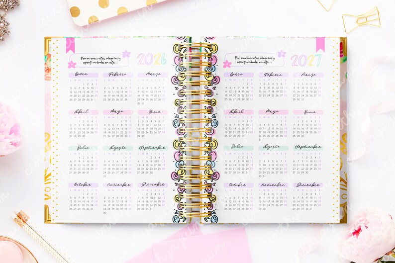 May include: Open planner with 2026 and 2027 calendars. The pages are white with pastel-colored month headings and gold spiral binding. The planner has a floral design on the edges and includes text in Spanish.