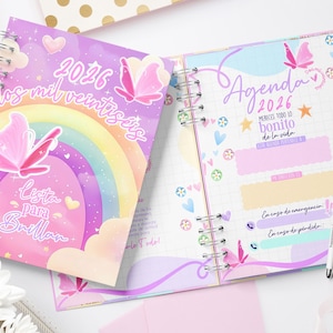 Puede incluir: Agenda 2026 con espiral, con una colorida portada que presenta un arcoíris, mariposas y el texto "Lista Para Brillar". Las páginas interiores muestran un diseño pastel con espacio para notas.