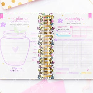 May include: Open planner with a gold spiral binding, featuring pink, purple, and gold accents. The pages include handwritten text and illustrations, including a jar and flowers. The planner is designed for organization and goal setting.