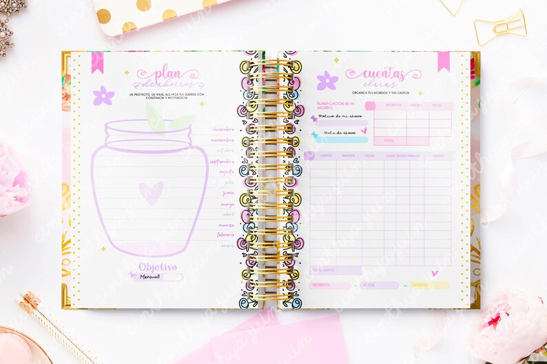 May include: Open planner with a gold spiral binding, featuring pink, purple, and gold accents. The pages include handwritten text and illustrations, including a jar and flowers. The planner is designed for organization and goal setting.