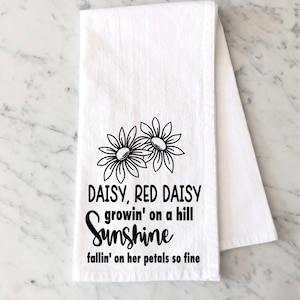 May include: White kitchen towel with black daisy flower illustrations and the text "DAISY, RED DAISY growin' on a hill Sunshine fallin' on her petals so fine". The towel is folded and has a marble background.
