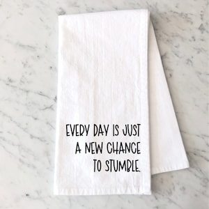 Op de afbeelding: Witte keukendoek met de tekst "EVERY DAY IS JUST A NEW CHANCE TO STUMBLE" in het zwart. De handdoek is gevouwen en ligt op een marmeren oppervlak. De tekst is in een vetgedrukte, schreefloze letter.