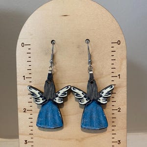 May include: A pair of angel-shaped earrings. Each earring features a blue dress, gray hair, and white and black wings. The earrings hang from silver-colored hooks. The earrings are displayed on a wooden stand with inch measurements.