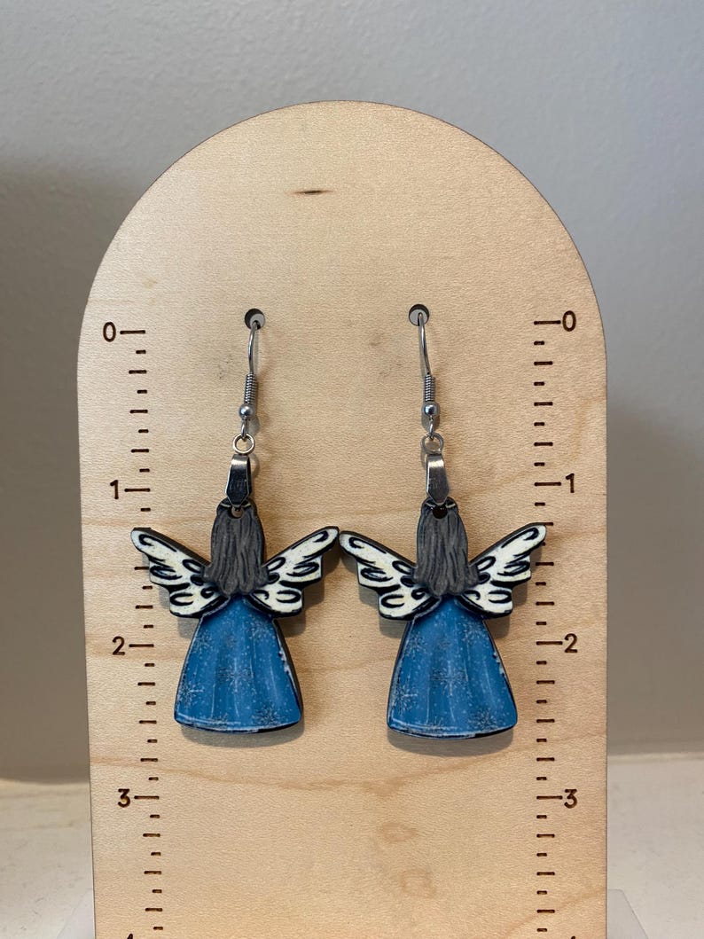 May include: A pair of angel-shaped earrings. Each earring features a blue dress, gray hair, and white and black wings. The earrings hang from silver-colored hooks. The earrings are displayed on a wooden stand with inch measurements.