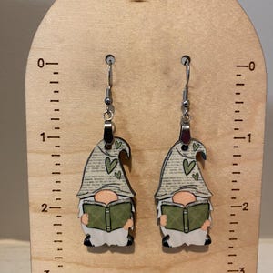 May include: A pair of gnome earrings. Each earring features a gnome reading a green book, with a gray hat adorned with green hearts. The earrings are suspended from silver-colored hooks and are displayed on a wooden stand.