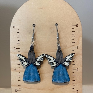 May include: A pair of angel-shaped earrings. The earrings feature a blue dress, white wings, and gray hair. The earrings are hanging from silver hooks. The earrings are displayed on a wooden ruler.