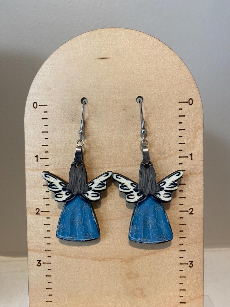 May include: A pair of angel-shaped earrings. The earrings feature a blue dress, white wings, and gray hair. The earrings are hanging from silver hooks. The earrings are displayed on a wooden ruler.