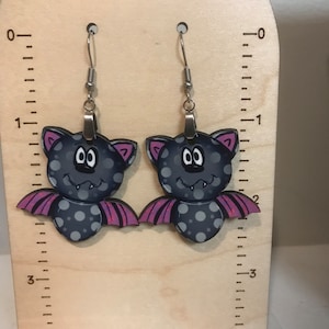 May include: A pair of black and white bat earrings with purple wings. The bats have white polka dots and a toothy grin.