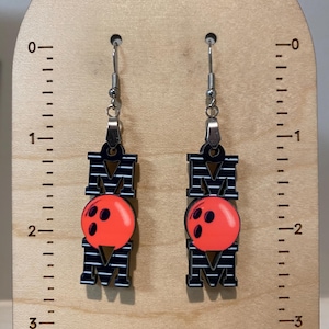 May include: A pair of black and white striped earrings with a red bowling ball charm. The earrings are hanging from silver hooks. The earrings are about 2 inches long.