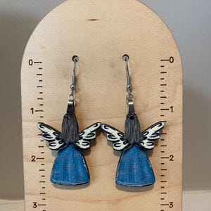 May include: A pair of angel-shaped earrings. Each earring features a blue dress, gray hair, and white and black wings. The earrings are suspended from silver-colored hooks. The earrings are displayed on a wooden stand with measurement markings.