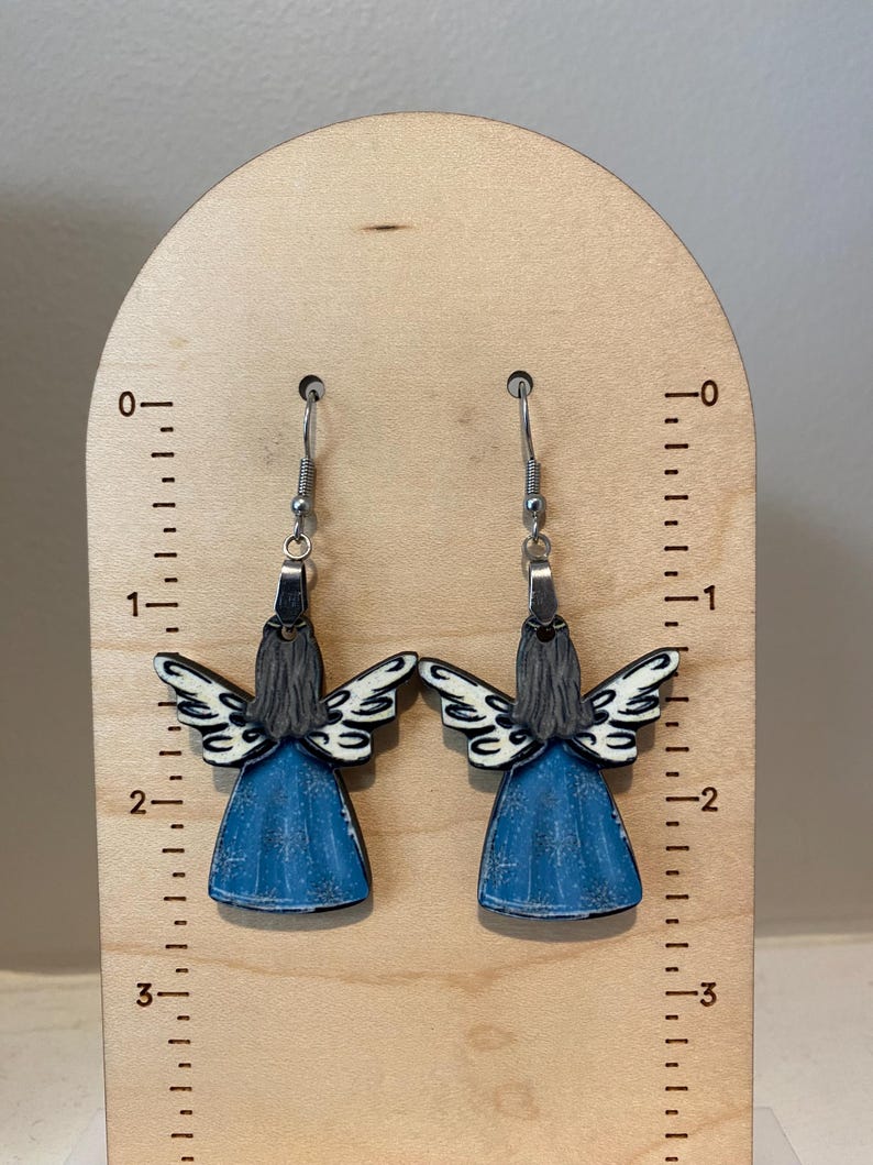 May include: A pair of angel-shaped earrings. Each earring features a blue dress, gray hair, and white and black wings. The earrings are suspended from silver-colored hooks. The earrings are displayed on a wooden stand with measurement markings.