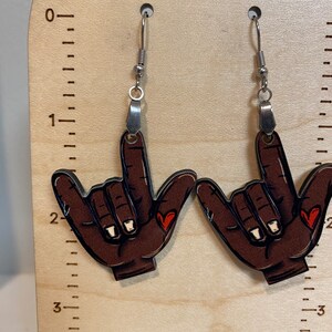 May include: A pair of brown wooden earrings shaped like a hand giving the shaka sign with a small red heart on each finger. The earrings are hanging from silver hooks.