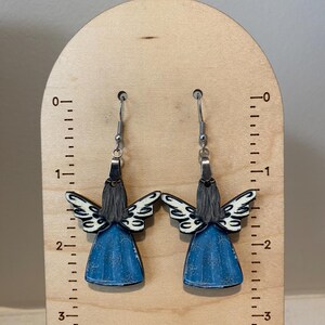 May include: A pair of angel earrings. Each earring features a blue dress, gray hair, and white and black wings. The earrings are hanging on a wooden measuring board.