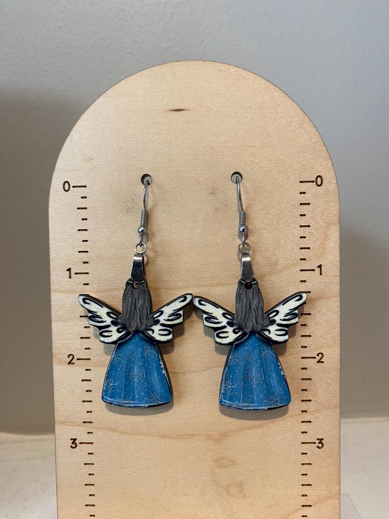 May include: A pair of angel earrings. Each earring features a blue dress, gray hair, and white and black wings. The earrings are hanging on a wooden measuring board.