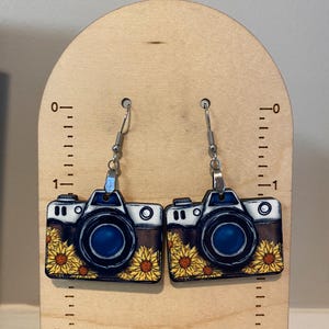 May include: A pair of camera-shaped earrings. The earrings feature a black and white camera design with a blue lens and a sunflower pattern. The earrings hang from silver-colored fish hook ear wires.