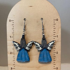 May include: A pair of angel-shaped earrings. Each earring features a blue dress, gray hair, and white wings with black detailing. The earrings are suspended from silver-colored hooks and are displayed on a wooden stand.