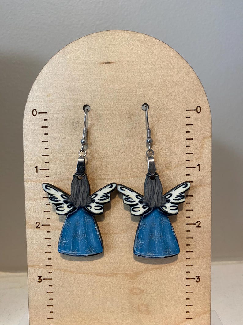 May include: A pair of angel-shaped earrings. Each earring features a blue dress, gray hair, and white wings with black detailing. The earrings are suspended from silver-colored hooks and are displayed on a wooden stand.