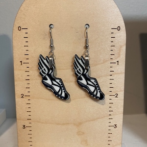 May include: A pair of black and white running shoe earrings with silver hooks. The earrings are displayed on a wooden stand with a ruler.