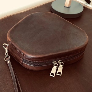 May include: A brown leather pouch with a zipper closure and a matching strap. The pouch is on a brown leather surface.