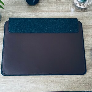 May include: A brown leather laptop sleeve with a grey felt top flap. The sleeve is on a wooden surface.