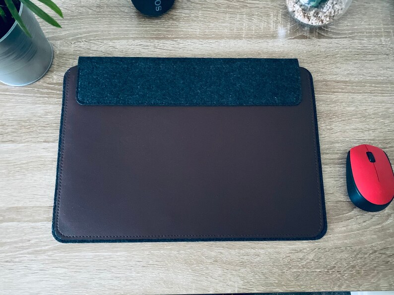 May include: A brown leather laptop sleeve with a grey felt top flap. The sleeve is on a wooden surface.