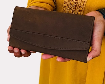 May include: A brown leather wallet with a flap closure. The wallet has a stitched design and is held in a person's hands.