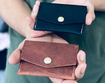 May include: Two leather wallets, one black and one brown, with a single button closure. Both wallets have white stitching around the edges.