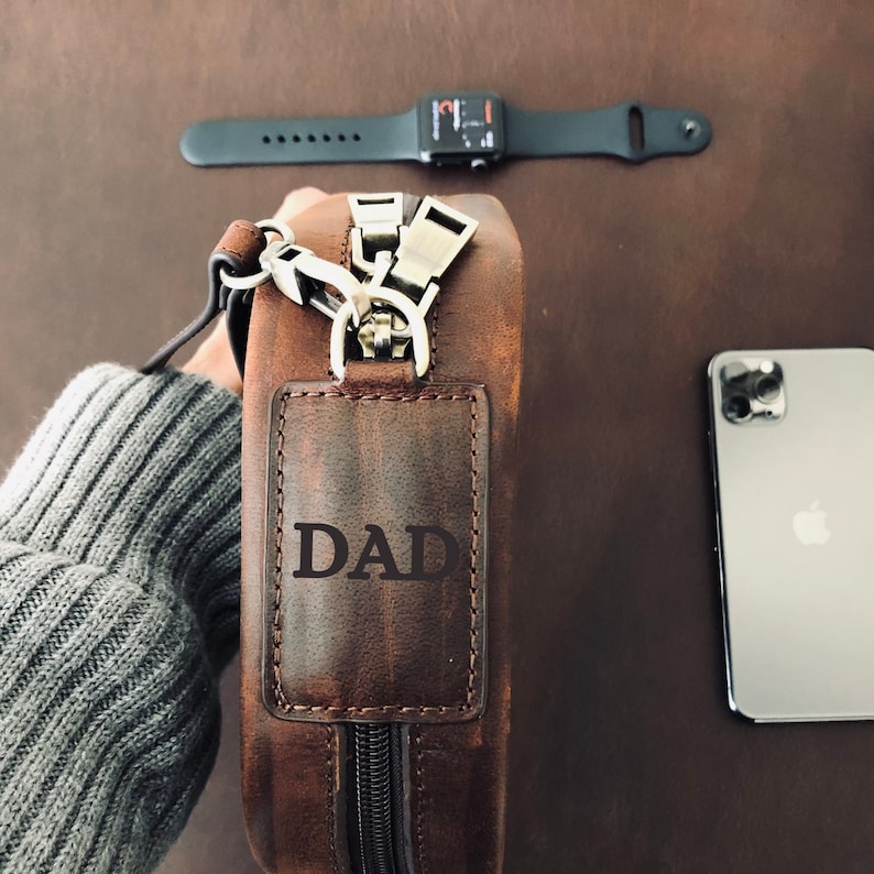 May include: A brown leather bag with a zipper closure and a strap. The bag has a small leather patch on the front with the word "DAD" embossed on it.