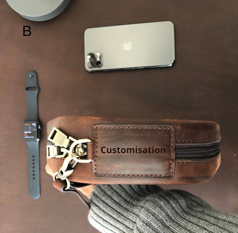 May include: A brown leather bag with a zipper closure and a brass buckle. The bag has a small rectangular patch on the front that says "Customisation".