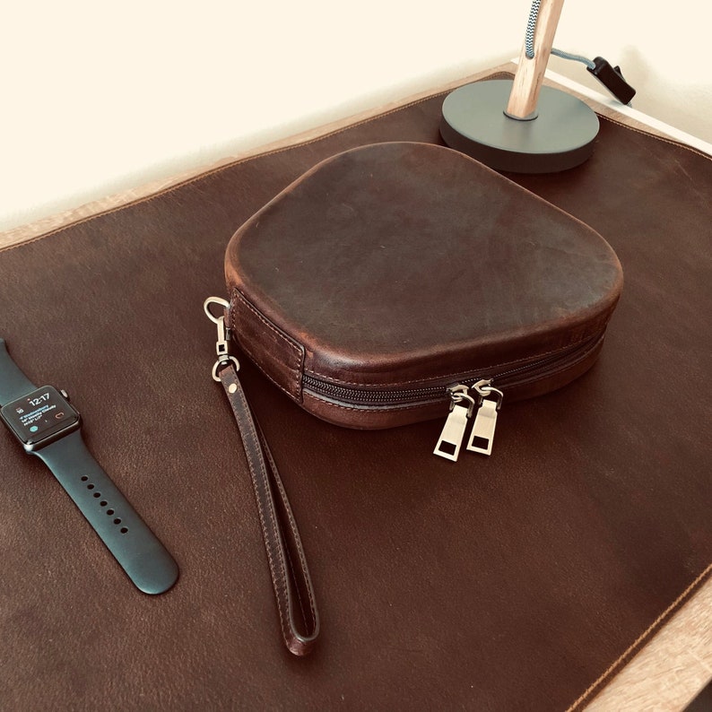 May include: A brown leather pouch with a zipper closure and a matching strap. The pouch is on a brown leather surface.