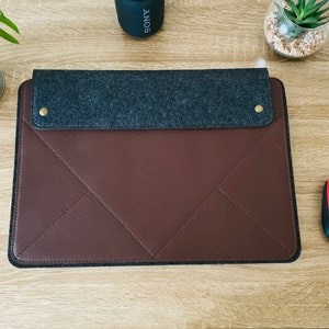 May include: A brown leather laptop sleeve with a grey felt flap and gold metal accents. The sleeve has a quilted design and is closed with a flap.
