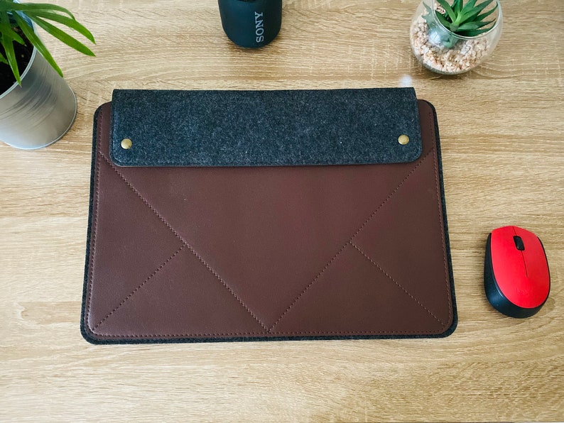 May include: A brown leather laptop sleeve with a grey felt flap and gold metal accents. The sleeve has a quilted design and is closed with a flap.