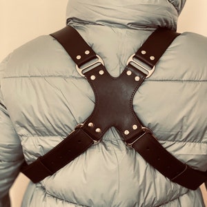 Puede incluir: Una correa de cámara de cuero negro con hebillas y remaches plateados. La correa está diseñada para llevarse cruzada sobre el pecho y la espalda.