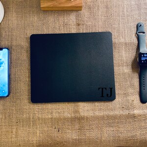 Pode incluir: Um tapete de rato preto com as iniciais "TJ" impressas em branco numa superfície de juta. Um Apple Watch preto com um mostrador branco também está na superfície.