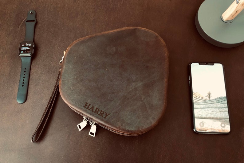 May include: A brown leather case with a zipper closure and the name "HARRY" embossed on the front. The case is sitting on a brown wooden surface next to a black Apple Watch and a black smartphone.