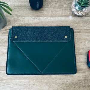 May include: A dark green leather laptop sleeve with a grey felt flap and two gold poppers. The sleeve is on a wooden surface.