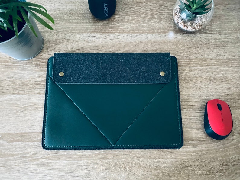 May include: A dark green leather laptop sleeve with a grey felt flap and two gold poppers. The sleeve is on a wooden surface.