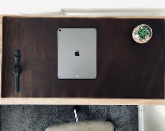 Può includere: Un tappetino da scrivania in pelle marrone con un bordo cucito. Un tablet computer argentato e uno smartwatch nero sono sul tappetino. Una piccola pianta in vaso si trova sulla scrivania a destra del tappetino.