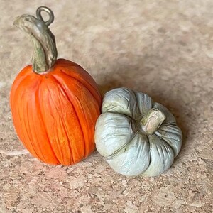 May include: Two miniature pumpkins. One is bright orange with a green stem and a metal loop. The other is a small, light green pumpkin with a textured surface. These decorative pumpkins are perfect for fall-themed crafts.