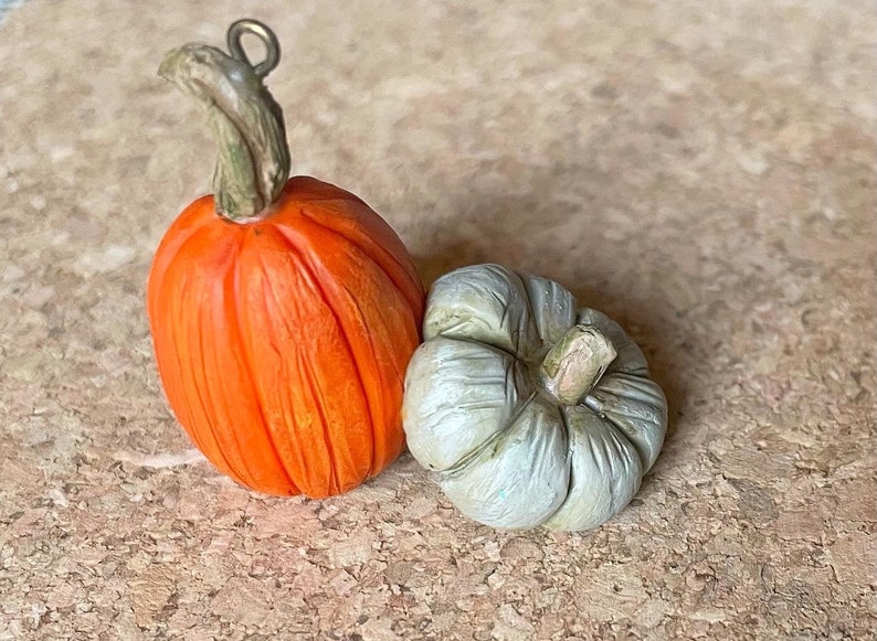 May include: Two miniature pumpkins. One is bright orange with a green stem and a metal loop. The other is a small, light green pumpkin with a textured surface. These decorative pumpkins are perfect for fall-themed crafts.