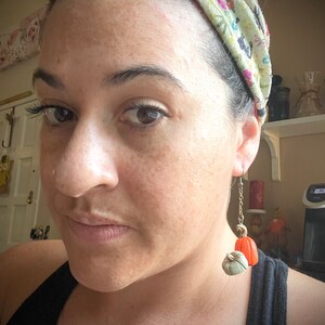 May include: A close-up shot of a person wearing pumpkin-shaped earrings. The earrings feature a small, light green pumpkin and a larger orange pumpkin, suspended from a gold-colored chain. The person is wearing a black tank top and a floral headband.